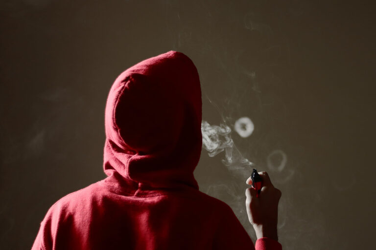 A hooded man blows a ring from his vape pen